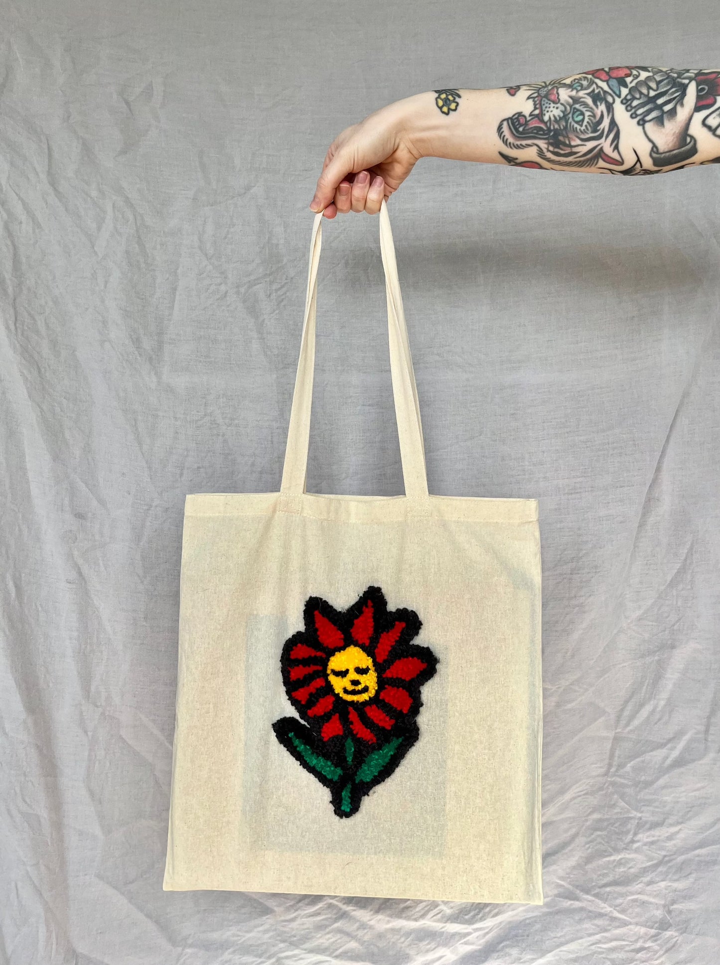 A hand holds a beige tote bag featuring a red and yellow punch needled flower with a green stem and two leaves against a grey background.