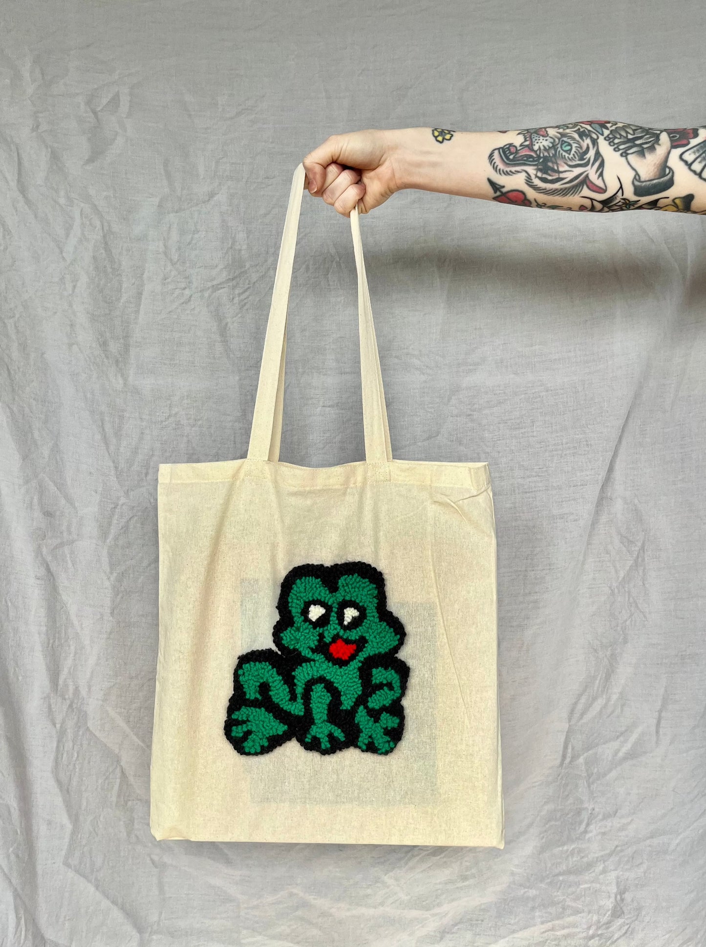 A hand holds a beige tote bag featuring a green punch needled frog with a red mouth against a grey background.
