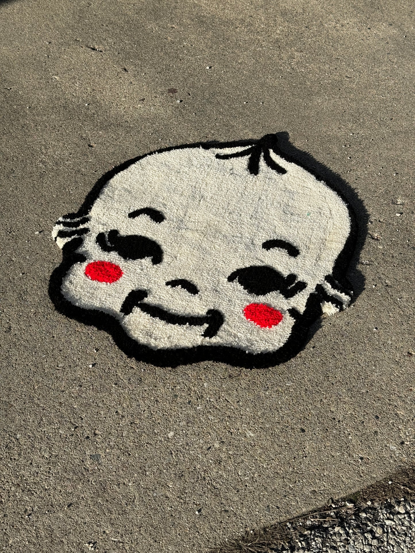 A tufted rug of a kewpie face with prominent red cheeks and expressive black features, set against a grey background.