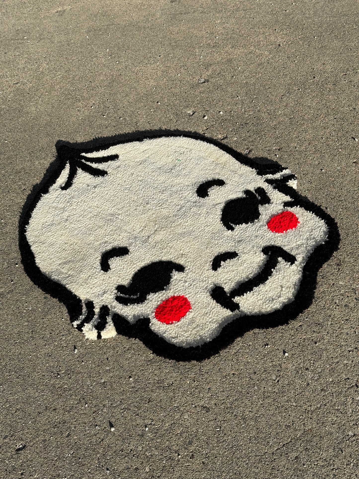 A tufted rug of a kewpie face with prominent red cheeks and expressive black features, set against a grey background.
