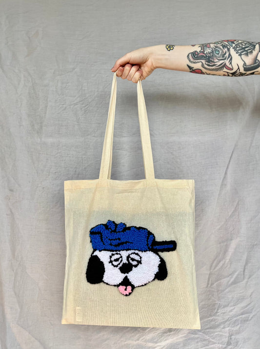     - A hand holds a beige tote bag featuring a white and black punch needled dog wearing a blue cap against a grey background.