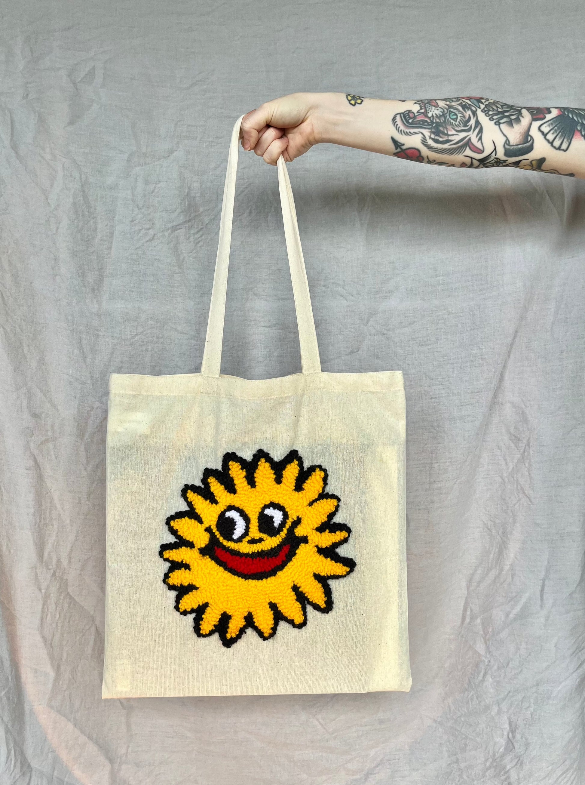 A hand holds a beige tote bag featuring a yellow punch needled sun with a smiling face with a bright red mouth against a grey background.