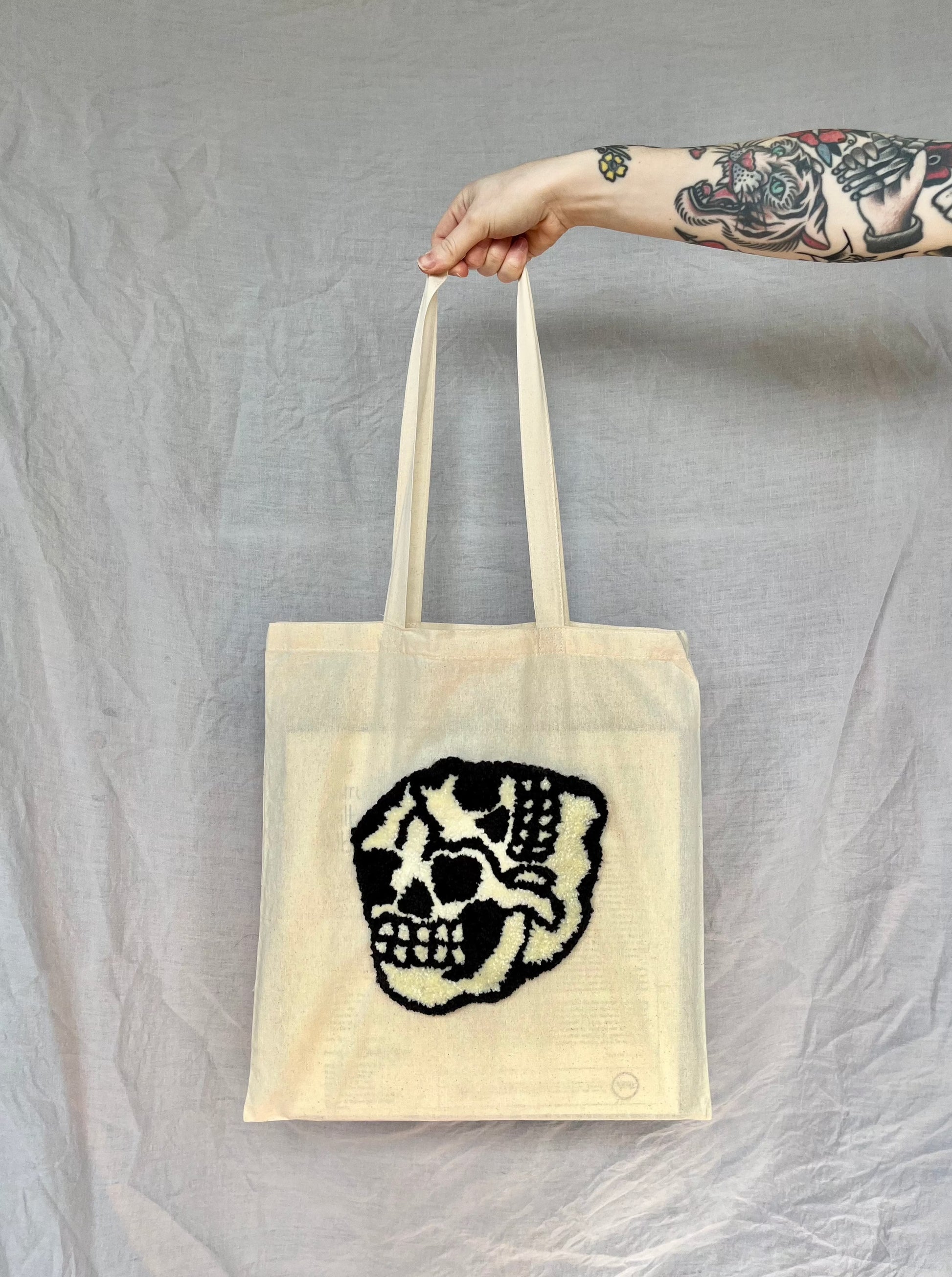 A hand holds a beige tote bag featuring a back and cream punch needled three-eyes skull against a grey background.