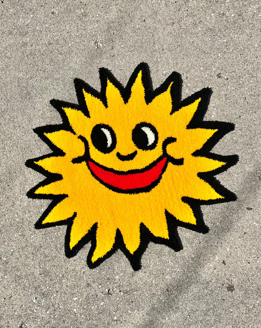 - A yellow tufted sun rug with a smiling face with a bright red mouth is shown against a grey background.