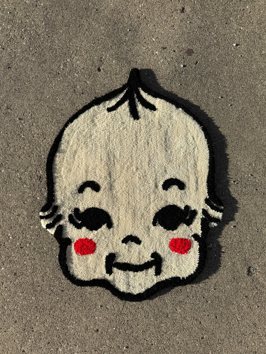 A tufted rug of a kewpie face with prominent red cheeks and expressive black features, set against a grey background.