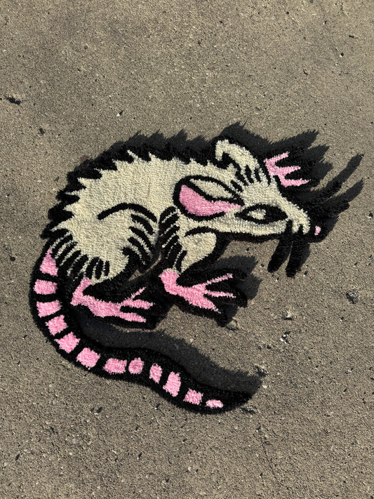 Black and white tufted rug of a rat with pink highlights and expressive eyes on a textured concrete surface.